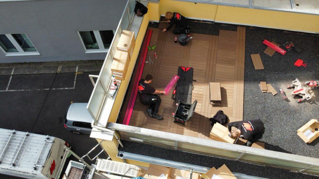 Draufsicht auf ein Team, das modulare Holz-/WPC‑Dielen auf der Dachterrasse verlegt; schwarze Tragschicht, Werkzeug und Materialkisten liegen bereit – Arbeitsschritt Belagsmontage zur schnellen Wiedervermietung.