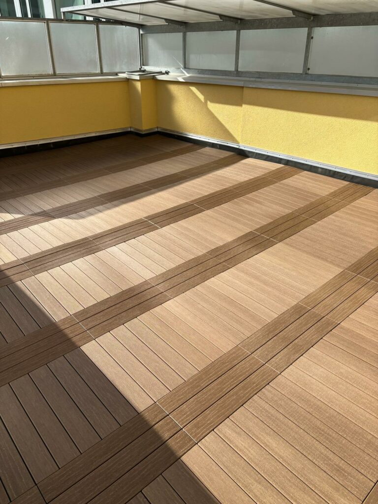Sonnige Dachterrasse nach der Sanierung: sauber verlegte Holz-/WPC‑Dielen in Bahnenoptik, aufgeräumte Rand‑ und Sockelanschlüsse, Glasgeländer – vermietungsbereit.