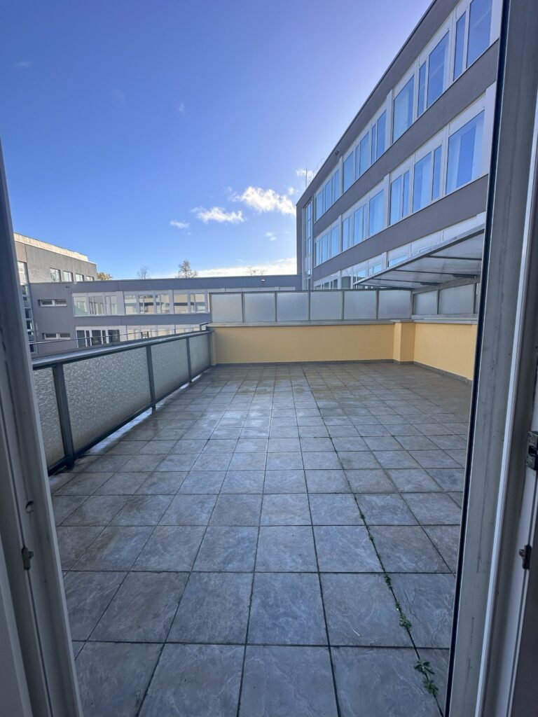 Weitwinkel auf die Dachterrasse: abgenutzte, graue Fliesen mit Bewuchs in den Fugen; Glasgeländer und gelbe Attika – Zustand vor der Sanierung.