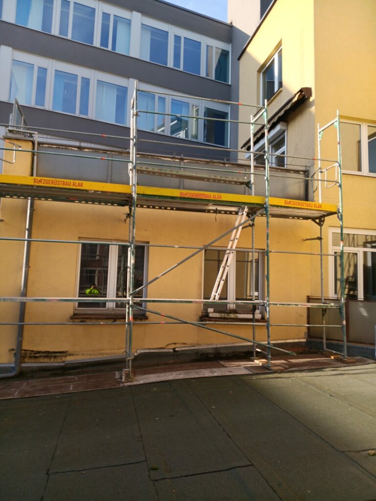 Entlang der gelben Hauswand steht ein Fassadengerüst; die Dachterrasse ist für Arbeiten an Brüstung, Anschlüssen und Abdichtung vorbereitet – während der Sanierung.