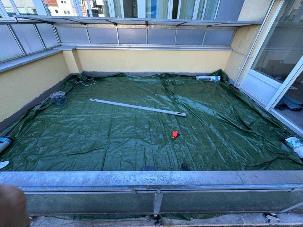 Blick von der Brüstung: grüne Plane schützt die frisch bearbeitete Fläche; Richtlatte, Eimer und Material am Rand – Vorbereitung für den nächsten Schritt des Bodenaufbaus.