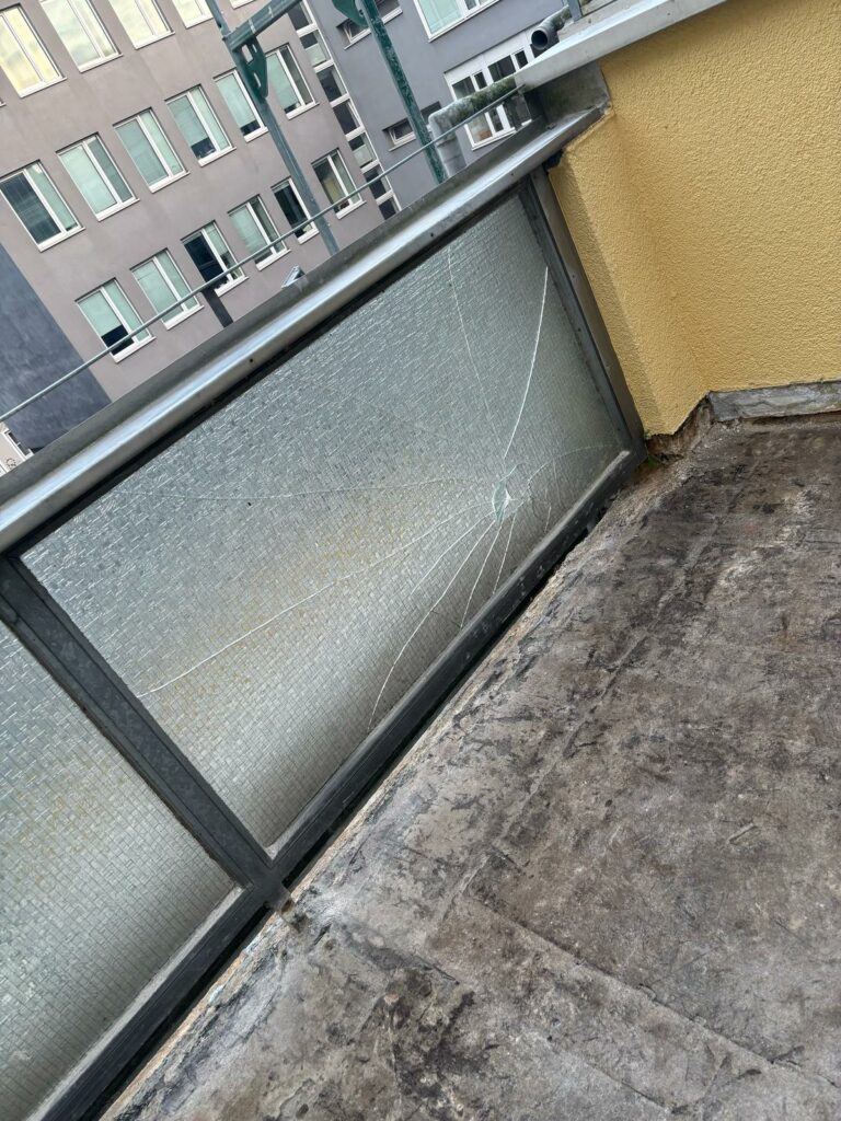 Nahaufnahme an der Glasbrüstung mit deutlich gerissenem Feld; Terrassenrand und Anschlussbereiche freigelegt und vorbereitet für Abdichtungs‑/Belagsarbeiten.