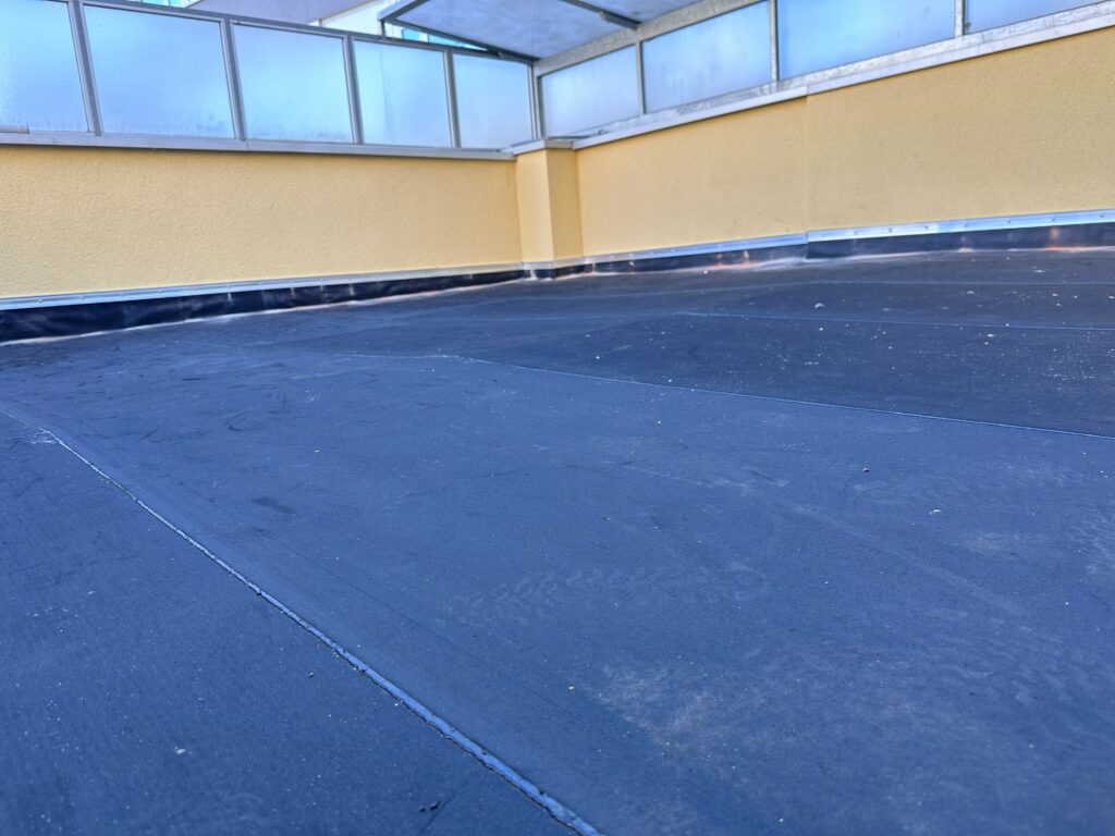 Niedrige Perspektive auf die Terrasse: durchgehende schwarze Abdichtung, hochgeführte Sockel‑/Wandanschlüsse; Glasbrüstung und gelbe Fassade im Hintergrund – Arbeitsschritt Abdichtung.